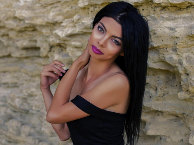DaisyKyra mit langen schwarzen Haaren, lila Lippenstift und einem schwarzen Top mit Schulterauschnitt, steht an einer Felswand und berührt ihren Hals mit einer Hand. Ihre Brüste sind sichtbar, sie sieht sexy aus..
