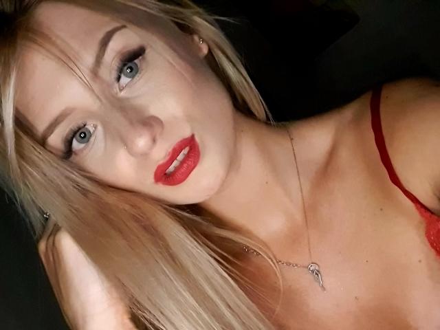 LenaaDoll with blonde hair, red lipstick, and a red lace bra, has blue eyes, a silver necklace, and a slight smile, looking at the camera.