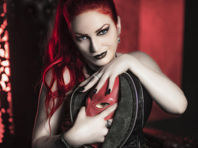 ZiaVyxen with red hair, black lipstick, and dark eye makeup, holding a red mask over her tits, wearing a black leather jacket, in a red-lit, dark room.