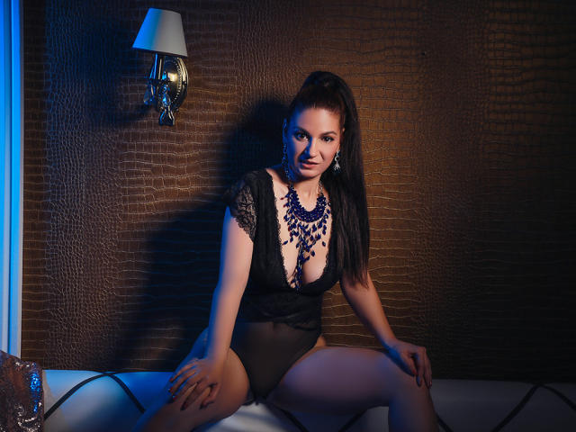 TenderLoveX with dark brown hair in a high ponytail, wearing a black lace bodysuit, blue necklace, sitting with legs apart, blue and warm light, crocodile textured wall, wall sconce.
