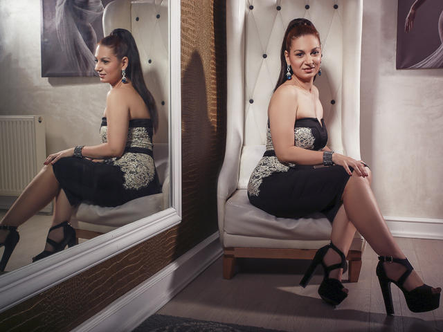 TenderLoveX sitting on a white, tufted chair, wearing a black dress with silver lace, black high heels, and earrings, with her ass and reflection in the mirror.