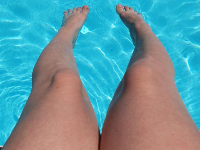 Die Beine von AngryGirl69, gebräunt und leicht muskulös, sind in einem leuchtend blauen Pool versunken, die Zehen zeigen nach oben, und man sieht kleine Wellen im Wasser, was eine entspannte, sommerliche Atmosphäre vermittelt..