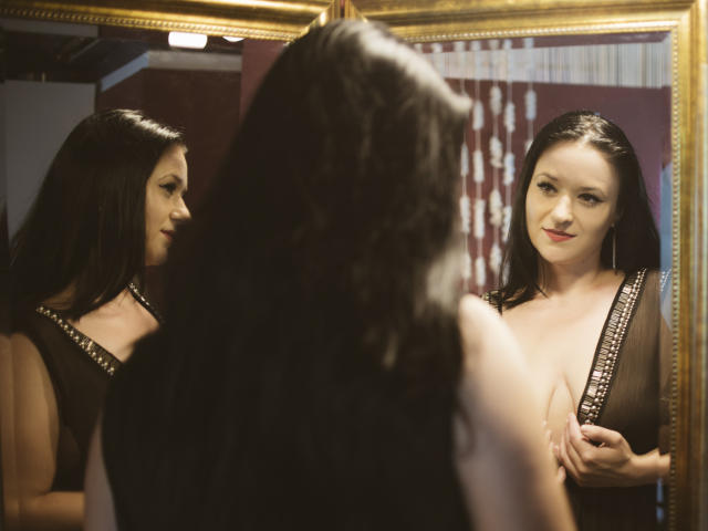 SensualCrissa with dark hair, wearing a black sheer dress with silver studs, standing in front of a mirror, gently touching her tits, smiling subtly, reflecting her face.