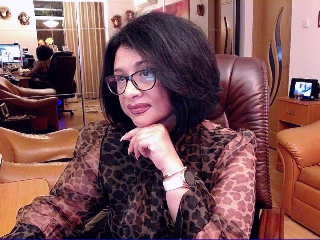 ClassybutNaughty with black hair, glasses, and lipstick, wearing a leopard-print shirt, sits in a brown leather chair, resting her chin on her hand, looking at the camera.