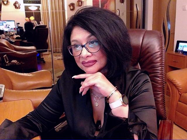 ClassybutNaughty with black hair, glasses, and lipstick, wearing a black top, showing her cleavage, pointing with her finger, sitting in a brown leather chair, smiling.