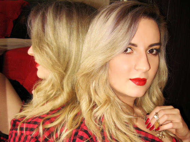 MoonBlossom with blonde hair, red lipstick, and red nails, looking at a mirror, wearing a red and black plaid shirt, holding a ring, reflected in a mirror.