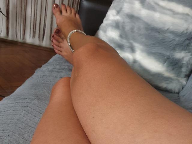 SexyCoco's legs crossed on a bed, showing tan skin, painted red toenails, and a silver ankle bracelet. Her ass is slightly visible.