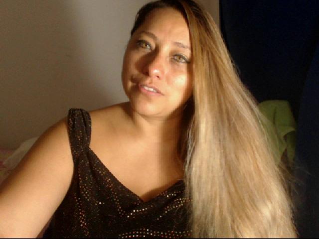 LovelyDiana69, con lunghi capelli biondi, occhi marroni e un viso leggermente paffuto, indossa una maglietta nera, scintillante e senza maniche, e guarda la fotocamera con un sorriso appena accennato..