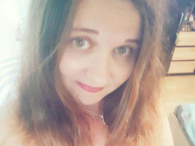 ChaudeBella69 ha gli occhi verde chiaro, i capelli castano ondulati e un leggero sorriso con rossetto rosso. Indossa una sottile collana. L'immagine è leggermente sfocata..