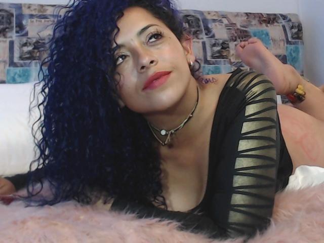 ScarlettBigAss with blue curly hair, black and gold striped top, red lipstick, necklace, lying on pink bed, feet up, looking at camera with a slight smile.