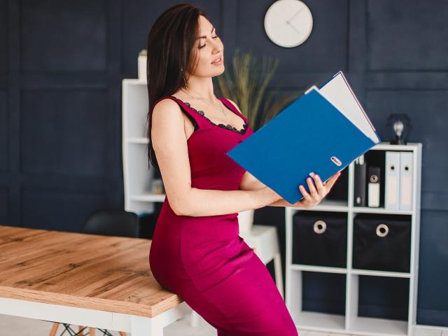 TellySabe, con cabello largo y castaño, vestida con un vestido rojo ajustado, sosteniendo un libro azul, sentada en una mesa de madera, mirando hacia abajo con una ligera sonrisa, mostrando sus senos..