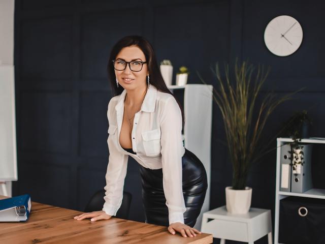 TellySabe con gli occhiali, una camicia bianca, pantaloni di pelle nera e un seno prosperoso, appoggiata a un tavolo di legno, sorridente, con capelli scuri, pareti bianche, un orologio, una pianta e una mensola sullo sfondo..