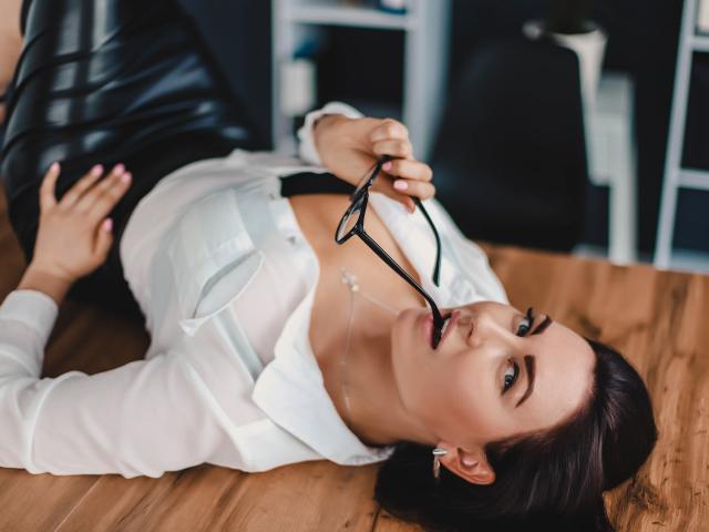 Les seins de TellySabe sont visibles sous une chemise blanche, déboutonnée, alors qu'elle est allongée sur une table en bois, tenant des lunettes noires dans sa bouche, et portant un pantalon en cuir noir..
