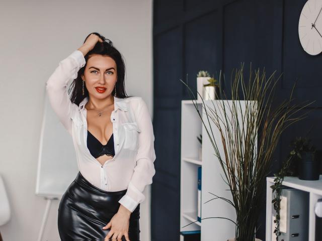 TellySabe with black lace bra, white shirt, and black leather skirt, hand in hair, red lipstick, and a confident smile, standing in a modern room, showing her tits.