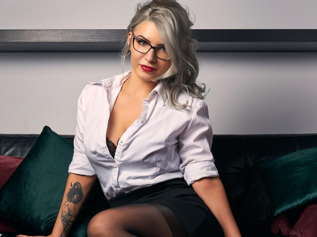 SandyKiss met grijs haar, een bril, rode lippenstift, een wit overhemd, een zwarte bh, een zwarte rok en een tatoeage op de rechterarm, zittend op een zwarte bank met groene en rode kussens..