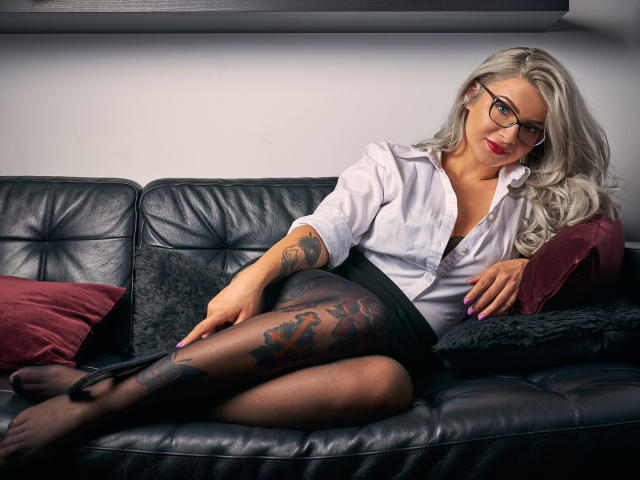 SandyKiss, avec des cheveux gris, des lunettes, une chemise blanche, une jupe noire et des collants transparents, montrant un tatouage sur sa cuisse droite, est allongée sur un canapé en cuir noir..