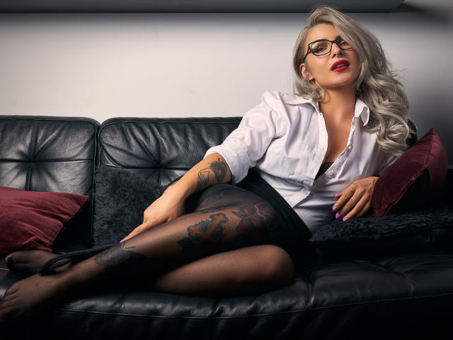 SandyKiss mit blonden Haaren, Brille, weißem Hemd, schwarzem Rock und transparenten schwarzen Strumpfhosen, die ein Tattoo an ihrem Oberschenkel zeigt, entspannt auf einem schwarzen Ledersofa mit roten Kissen..