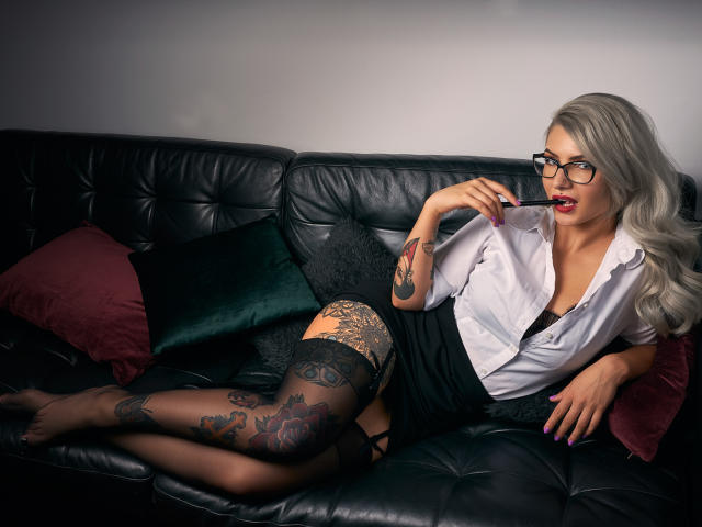SandyKiss with long gray hair, glasses, white shirt, black skirt, and lace thigh-highs, sucking a red lollipop, lying on a black leather couch, showing tattoos on legs.