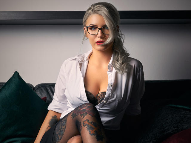 SandyKiss, avec des cheveux gris, des lunettes, une chemise blanche, un soutien-gorge en dentelle noire et des tatouages sur les jambes, assise sur des coussins vert foncé et noirs, regardant séduisamment l'appareil photo..
