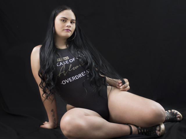 JassmineTS mit langen, schwarzen Haaren, die ein schwarzes Tanktop und einen hochgeschnittenen Body trägt, sitzt mit gespreizten Beinen, wobei ihr Hintern und ein Tattoo auf ihrem rechten Arm sichtbar sind..