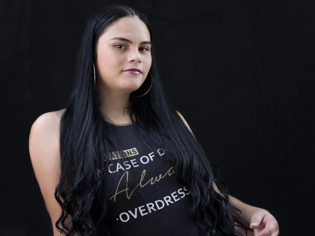 JassmineTS tem cabelos longos e pretos, veste uma blusa preta sem mangas, usa brincos grandes e batom roxo, e olha ligeiramente para o lado com uma expressão neutra..