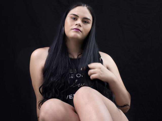 JassmineTS, com cabelos longos e pretos, batom roxo, vestindo uma blusa preta sem mangas, sentada com os joelhos levantados, a mão direita tocando o cabelo, fundo preto, olhando ligeiramente para baixo, expressão confiante..