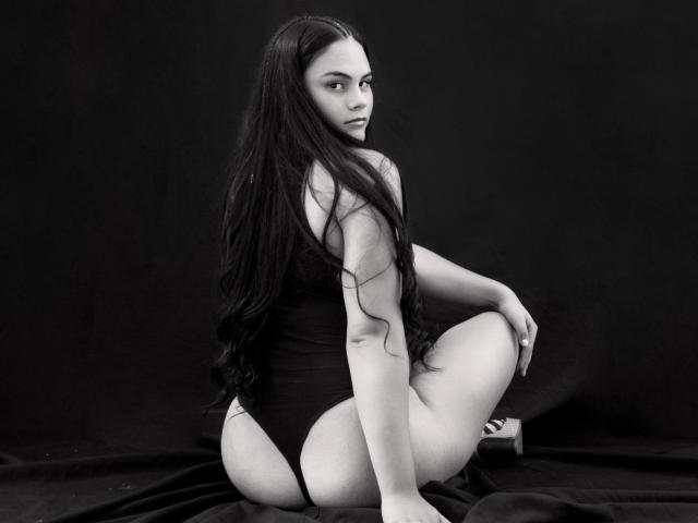 JassmineTS, con cabello largo y negro, sentada sobre una tela oscura, vestida con un traje de baño negro de una sola pieza, mostrando su trasero y mirando por encima del hombro con una expresión seria, en blanco y negro..