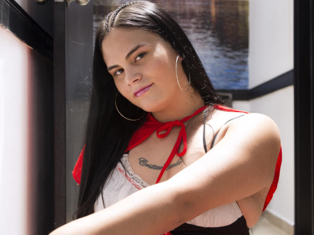 JassmineTS has long black hair, red halter top, hoop earrings, and a tattoo under her tits. She's smiling, with sunlit skin and pink lipstick. Her arms are crossed.
