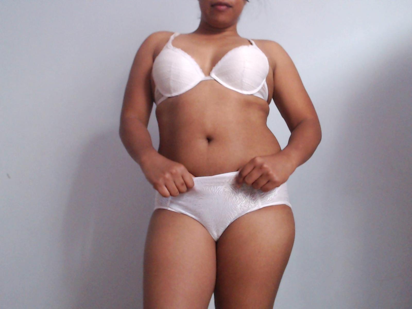 ShanniaXX met een medium bruine huid, draagt een witte bh en witte onderbroek, staat, met haar handen op haar taille, en laat haar borsten en vagina zien, in een eenvoudige, binnenomgeving..