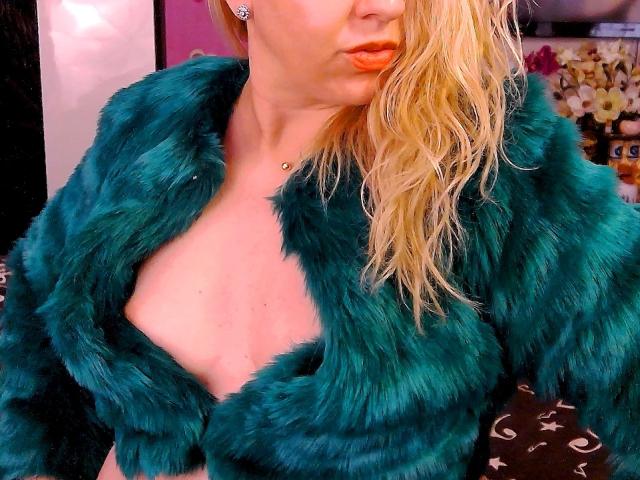 HotKrystall con i capelli biondi, labbra arancioni carnose e un cappotto di pelliccia color turchese, che mostra un po' il seno, indossa una piccola collana e orecchini in una foto sexy..