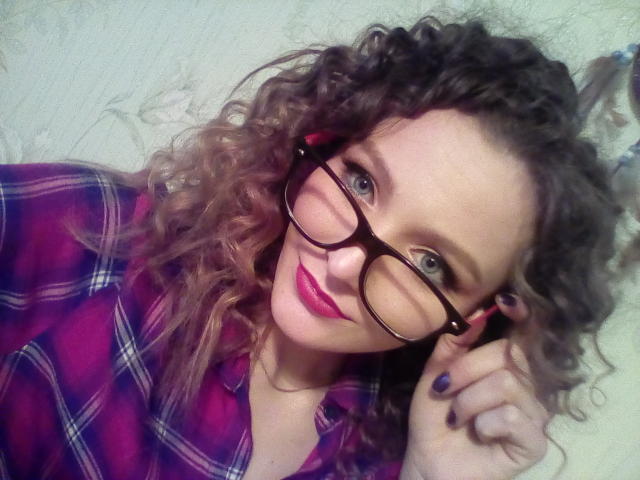 DannaShiny, avec ses cheveux bruns bouclés, portant une chemise à carreaux rouges, des lunettes noires, du rouge à lèvres violet et du vernis à ongles violet, souriant à la caméra, tenant des lunettes avec sa main droite..