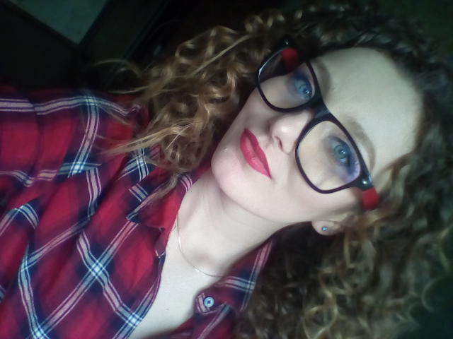 DannaShiny, avec ses cheveux roux-brun bouclés, porte une chemise à carreaux rouges et des lunettes noires. Elle a les yeux bleus, du rouge à lèvres rouge et un collier. Ses seins sont partiellement visibles..