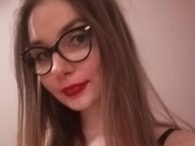 EllyBrune with long brown hair, wearing glasses, red lipstick, and black strap dress, showing a slight smile, looking directly at the camera.