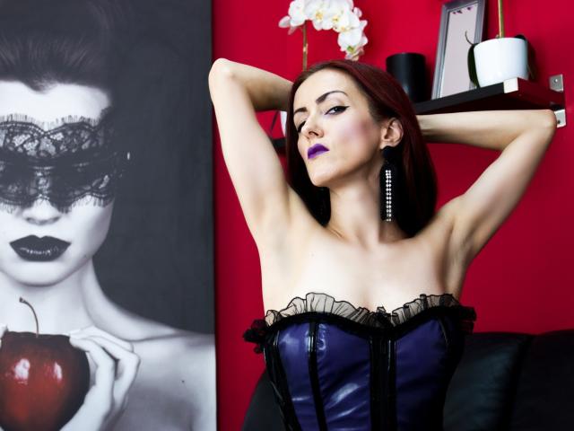ClaraDomme with purple lipstick, arms raised, wearing a black and purple corset, showing her tits, standing in front of a black and white portrait, red background.