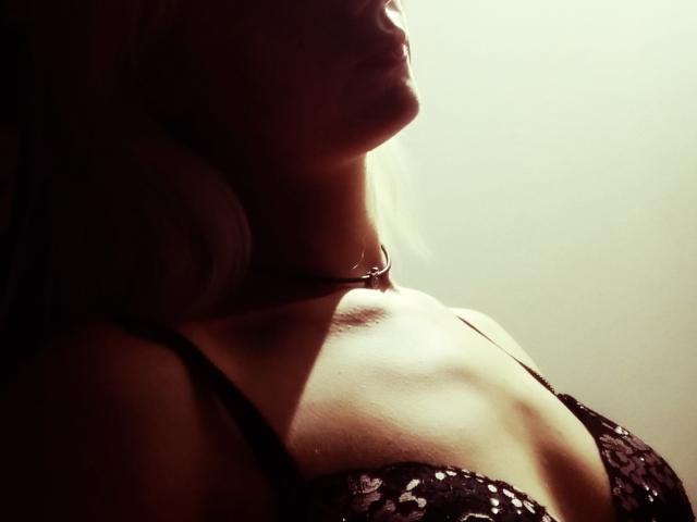 El perfil de SwissMiss, con un sujetador de encaje oscuro, mostrando sus pechos, su cuello delgado y su cabello rubio. Una luz suave resalta su clavícula y su pecho. Fotografía..