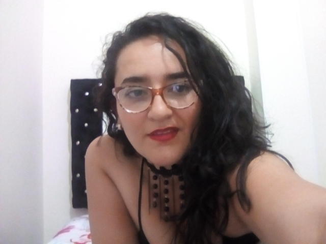 SaimaJaye69 with wavy black hair, glasses, red lipstick, and black choker with studs, showing bare shoulders and a black top, sitting on a bed.