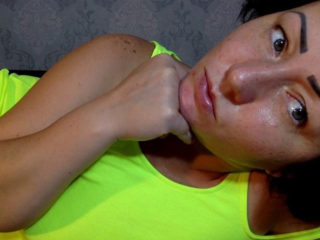 LoveJoySex con un'espressione seria, tenendosi il mento con la mano destra, indossa una canotta verde neon, mostrando il seno e la pelle lentigginosa, in una stanza buia..