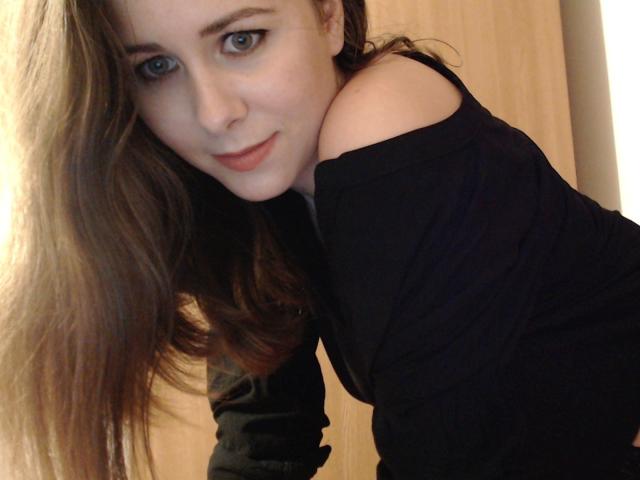 MissDonia met lang bruin haar, blauwe ogen en een lichte glimlach, draagt een zwarte top met een open schouder, leunt voorover en laat haar blote schouder en schattige gezicht zien..