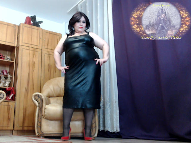 KaryQueen, plus-size woman with pale skin, black leather dress, black tights, red heels, standing, hands on hips, dark hair, red lipstick, wooden cabinets, beige chair, purple curtain.
