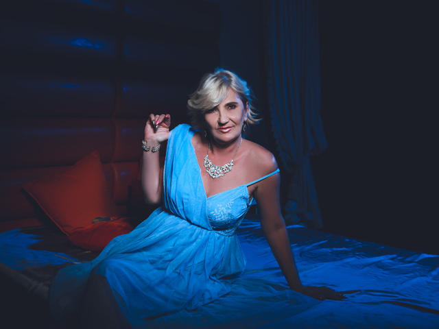 ExperiencedAlana with blonde hair, blue dress, off-shoulder, showing tits, sitting on bed, blue light, holding wrist, necklace, smiling, red pillow, dark room.