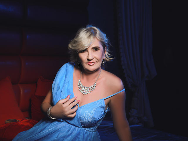 Alana, une femme expérimentée, avec des cheveux blonds courts, une robe bleue à épaules dénudées, laissant entrevoir sa poitrine, tenant la robe, portant un collier, assise sur un lit avec des coussins rouges, souriante, sur un fond sombre..
