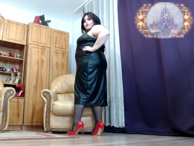 KaryQueen stands in a black leather dress, red high heels, black stockings, and red lipstick, with her hand on her hip, showing her curvy ass and large tits.