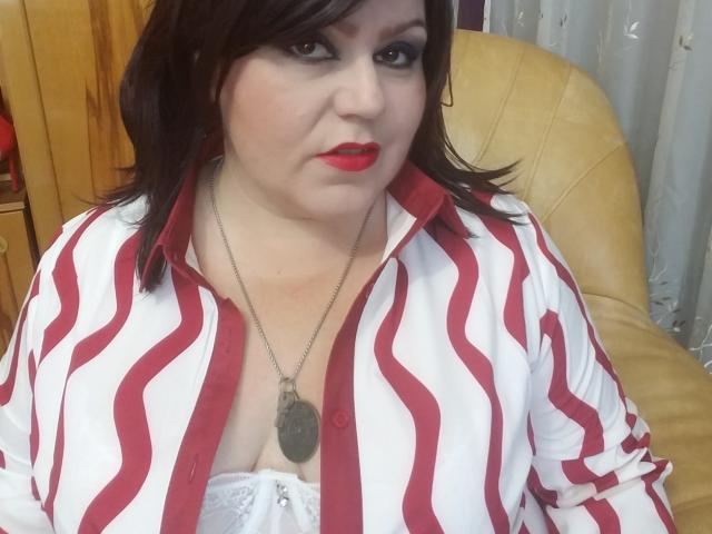 KaryQueen indossa una camicia a righe rosse e bianche, ha il rossetto rosso, i capelli neri, una collana e un reggiseno di pizzo bianco che mette in mostra il suo seno prosperoso. È seduta su una poltrona in pelle gialla..
