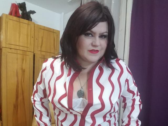 KaryQueen with wavy dark brown hair, red lipstick, and white shirt with red stripes, showing her large boobs, standing in a room with wooden cabinets and a purple curtain.