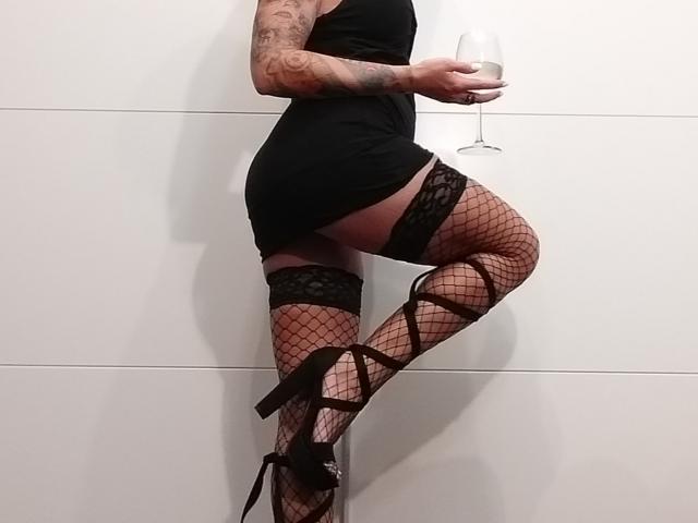 HotGirlVibe mit Tattoos, trägt ein schwarzes Kleid, ein Spitzenoberteil, hohe Strümpfe, Netzstrümpfe und High Heels, hält ein Weinglas, zeigt ihr Gesäß und ihre Beine..