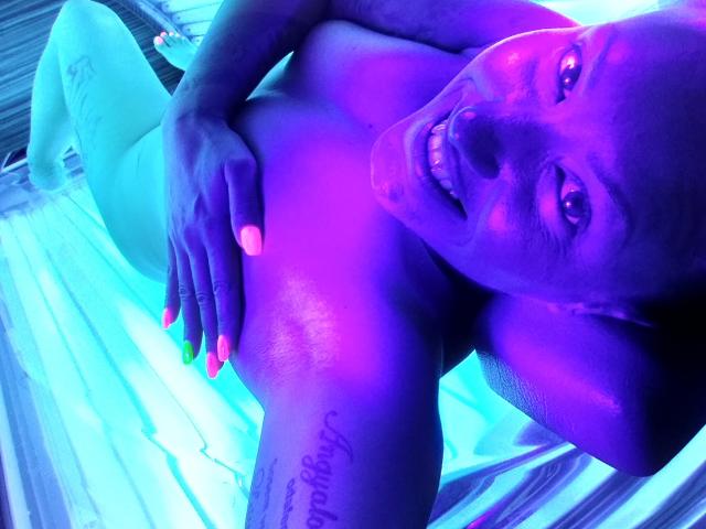 HotGirlVibe sdraiata su un lettino abbronzante con luce viola-blu, sorridente, calva con un tatuaggio sulla spalla destra, mano sul petto, unghie rosa e verde neon..