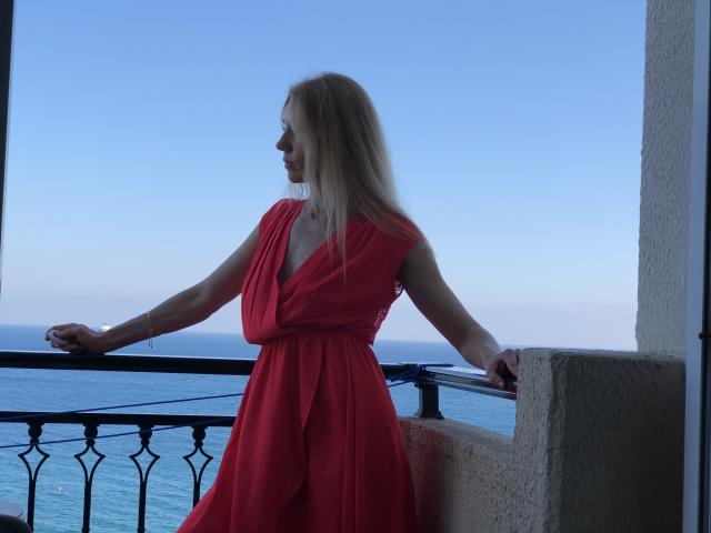 SugarLover in a red dress, standing with arms on a balcony, looking at the ocean. She has blonde hair, a slim build, and a slight smile.