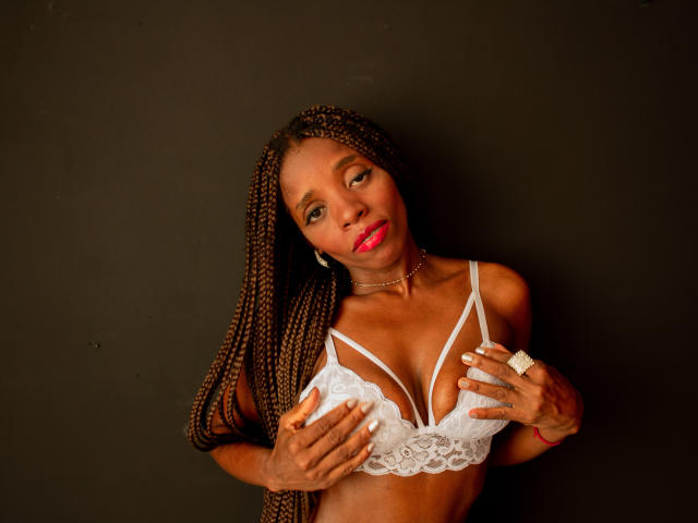 SandyChaudeX with dark skin, long braids, wearing white lace bra, hands on her tits, red lipstick, gold ring, black background, looking at camera with slight smile.