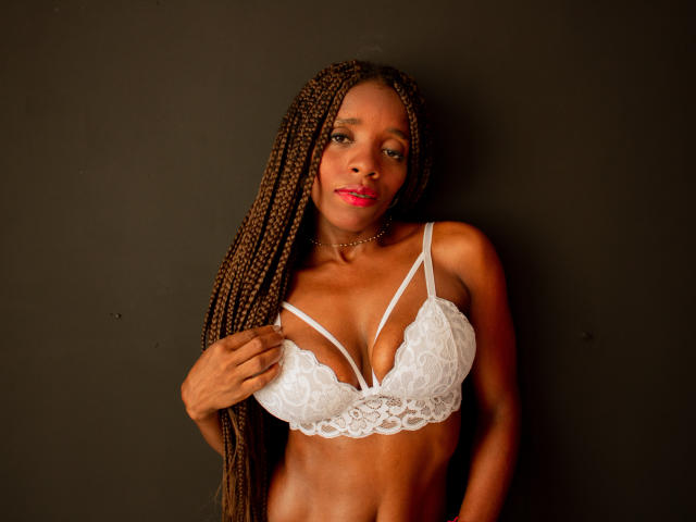 SandyChaudeX with long braids, wearing a white lace bra, holding it with one hand, showing her tits, standing against a dark background, red lipstick, confident expression.