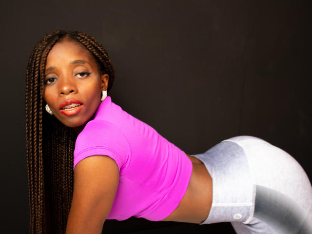 SandyChaudeX with long braided hair, pink crop top, and silver pants, on knees, butt up, looking over shoulder, smiling with red lipstick, white earrings, black background.
